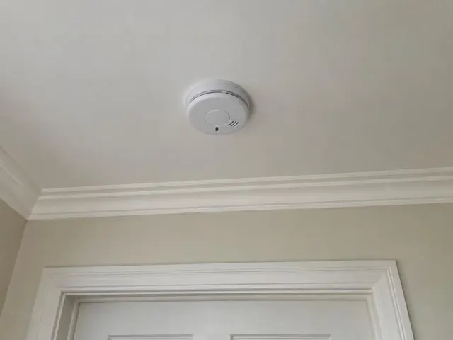White smoke alarm mounted on a ceiling in a beige room with cornice and door trim in Cooranbong